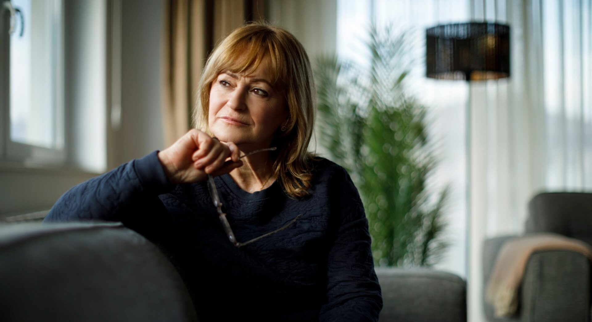 Vrouw met bril in hand zit op de bank en kijkt naar buiten